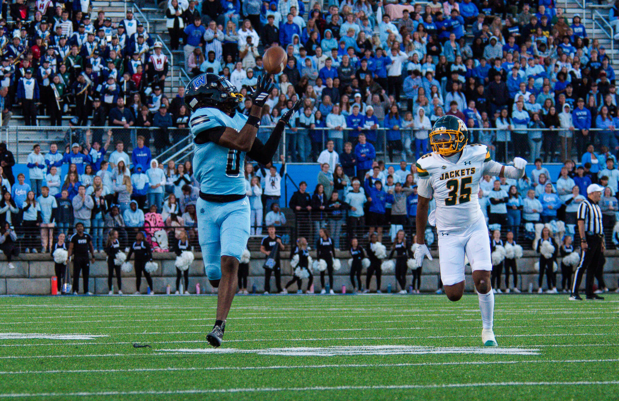 A football player in a light blue uniform jumps to catch the ball during a game, while an opposing player in a white and yellow uniform runs toward him. A large crowd of spectators fills the stadium seats in the background